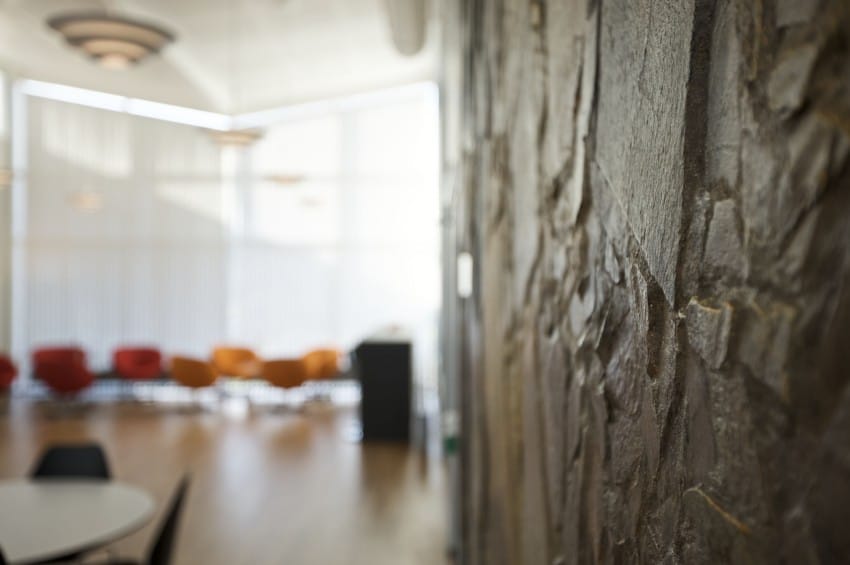 Nærbilde av en teksturert vegg i et lyst rom med moderne stoler og store vinduer i bakgrunnen. Foto: Nathan Lediard, Lediard Foto AS.