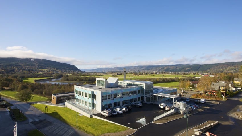 Moderne bygning med store vinduer omgitt av et naturskjønt landskap av jorder, åser og en svingete elv. Foto: Nathan Lediard, Lediard Foto AS.