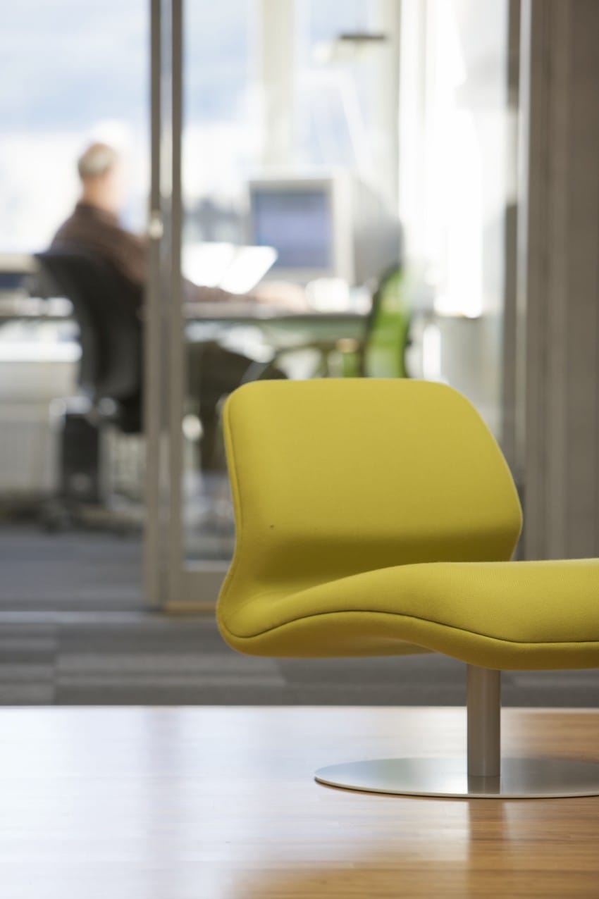 Gul stol i fokus på moderne kontor; uskarp bakgrunn viser person ved skrivebordet. Foto: Nathan Lediard, Lediard Foto AS.