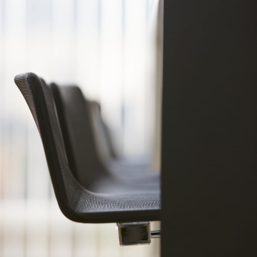 Nærbilde av svarte kontorstoler stilt opp ved siden av et bord, med uskarpe loddrette persienner i bakgrunnen. Foto: Nathan Lediard, Lediard Foto AS.