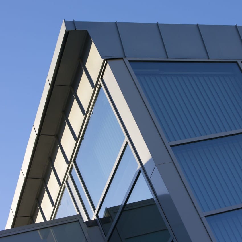 Moderne byggehjørne med glassfasade og metallpaneler mot en klar blå himmel. Foto: Nathan Lediard, Lediard Foto AS.