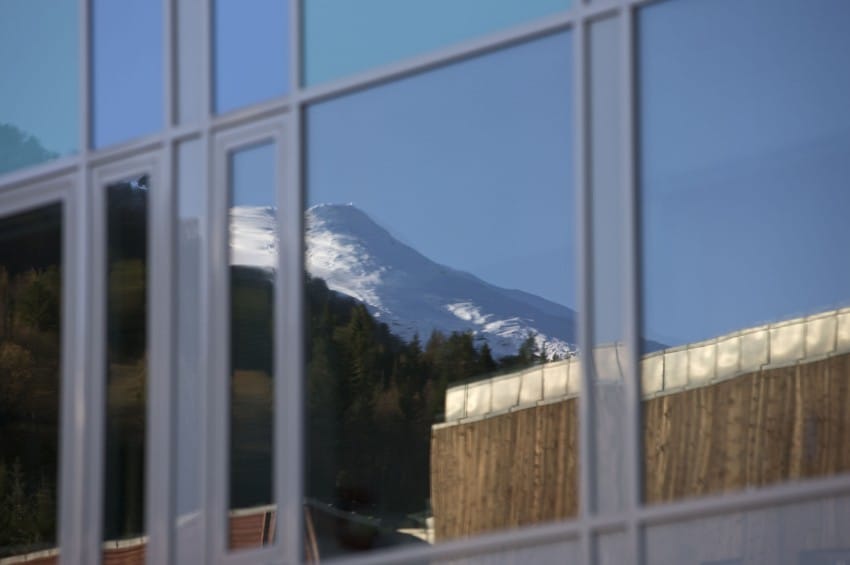 Refleksjon av et snødekt fjell og trær i glassvinduene til en bygning. Foto: Nathan Lediard, Lediard Foto AS.