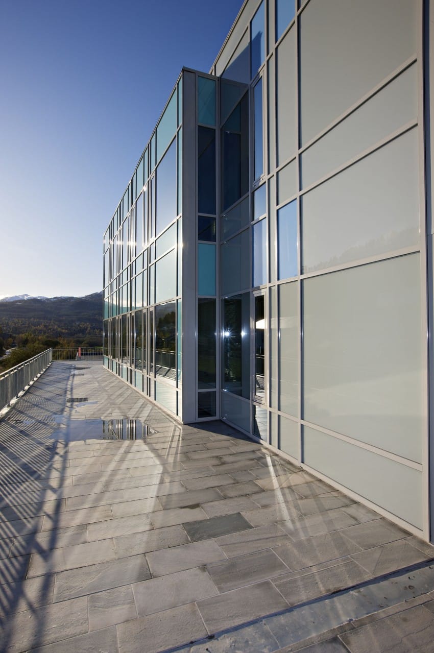 Moderne glassbygning med store vinduer som reflekterer himmelen, langs en solbelyst gangvei med steinflis, under en klarblå himmel. Foto: Nathan Lediard, Lediard Foto AS.
