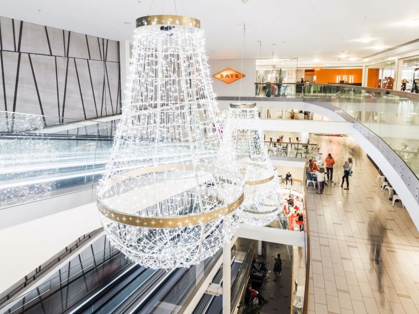 Sterkt opplyste lysekroner i et moderne kjøpesenter med flere nivåer og glassrekkverk, folk som går under. Foto: Nathan Lediard, Lediard Foto AS.