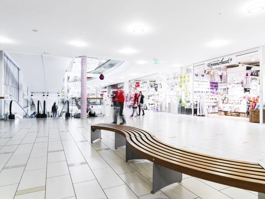 Romslig, moderne kjøpesenterinteriør med trebenker og butikker; folk som går i nærheten. Foto: Nathan Lediard, Lediard Foto AS.