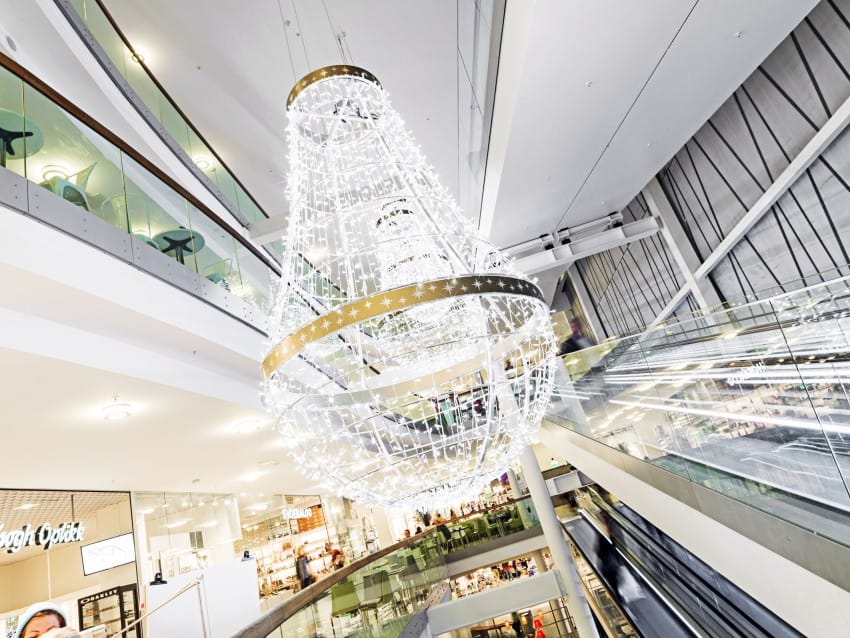 Stor opplyst lysekrone i et moderne kjøpesenteratrium med rulletrapper og butikker under. Foto: Nathan Lediard, Lediard Foto AS.