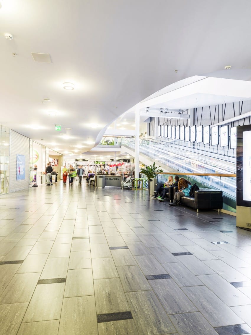 En kjøpesenterkorridor med folk som sitter på en sofa og går, rulletrapper til høyre. Foto: Nathan Lediard, Lediard Foto AS.