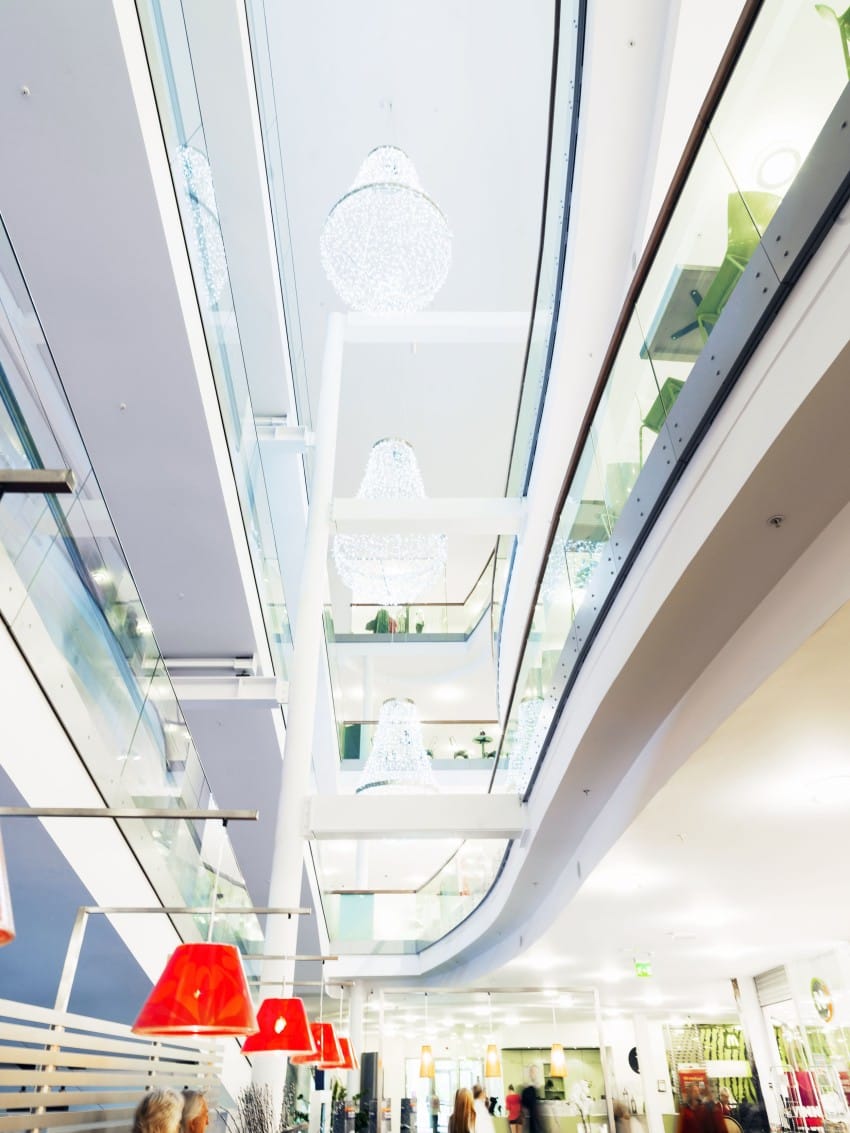 Moderne multi-etasjes kjøpesenterinteriør med glassrekkverk, store lysekroner, røde hengende lys og shoppere under. Foto: Nathan Lediard, Lediard Foto AS.