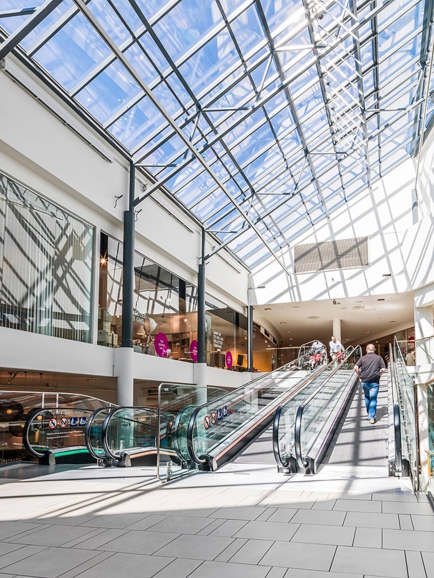 Moderne kjøpesenterinteriør med rulletrapper, glasstak og noen få mennesker som går. Foto: Nathan Lediard, Lediard Foto AS.