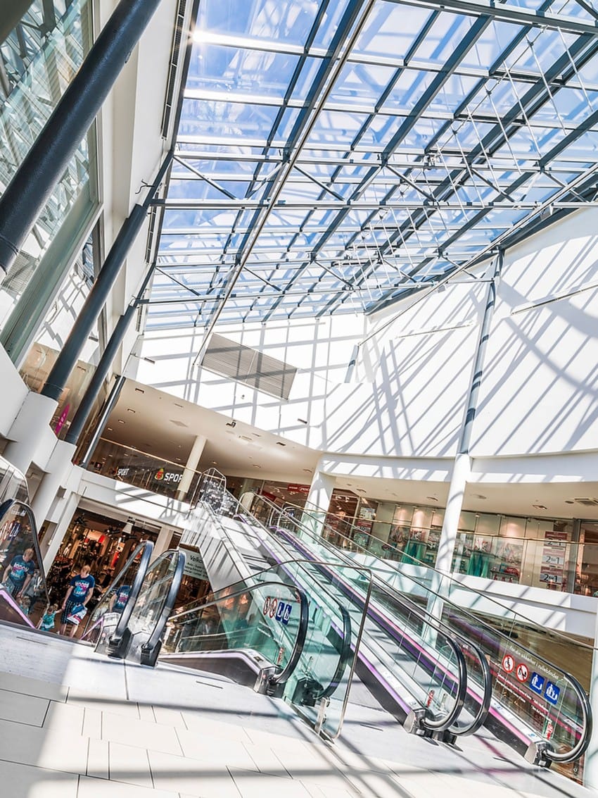 Kjøpesenterinteriør med glasstak, rulletrapper og folk som går. Sterkt sollys kaster skygger på gulvet. Foto: Nathan Lediard, Lediard Foto AS.