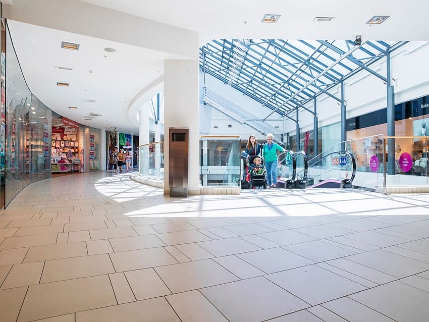En familie med barnevogn står på toppen av en rulletrapp i et lyst, romslig kjøpesenter. Foto: Nathan Lediard, Lediard Foto AS.