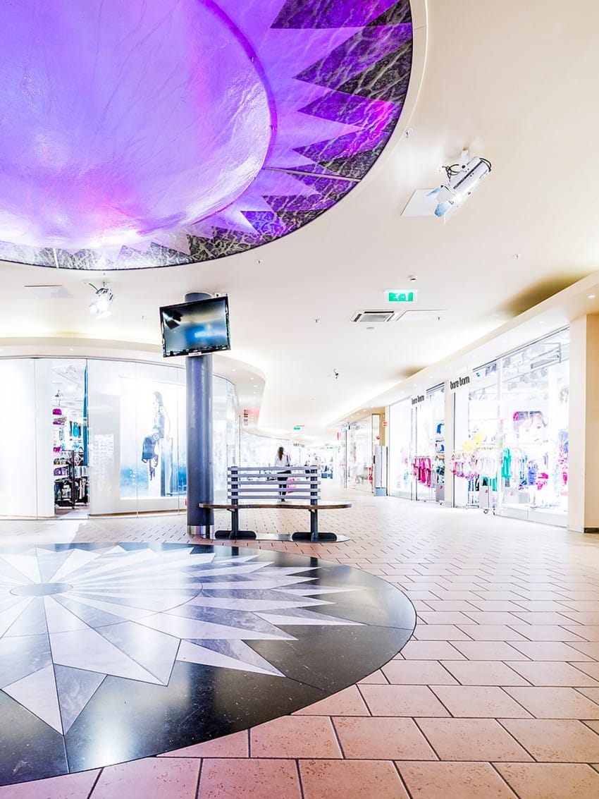 Moderne kjøpesenter med skarp belysning, lilla taksirkel og butikker med glassutstillinger. Foto: Nathan Lediard, Lediard Foto AS.