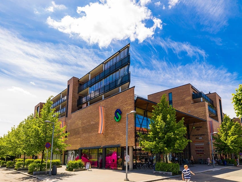 Moderne murbygning med butikker og leiligheter, omgitt av trær under en blå himmel med skyer. Foto: Nathan Lediard, Lediard Foto AS.