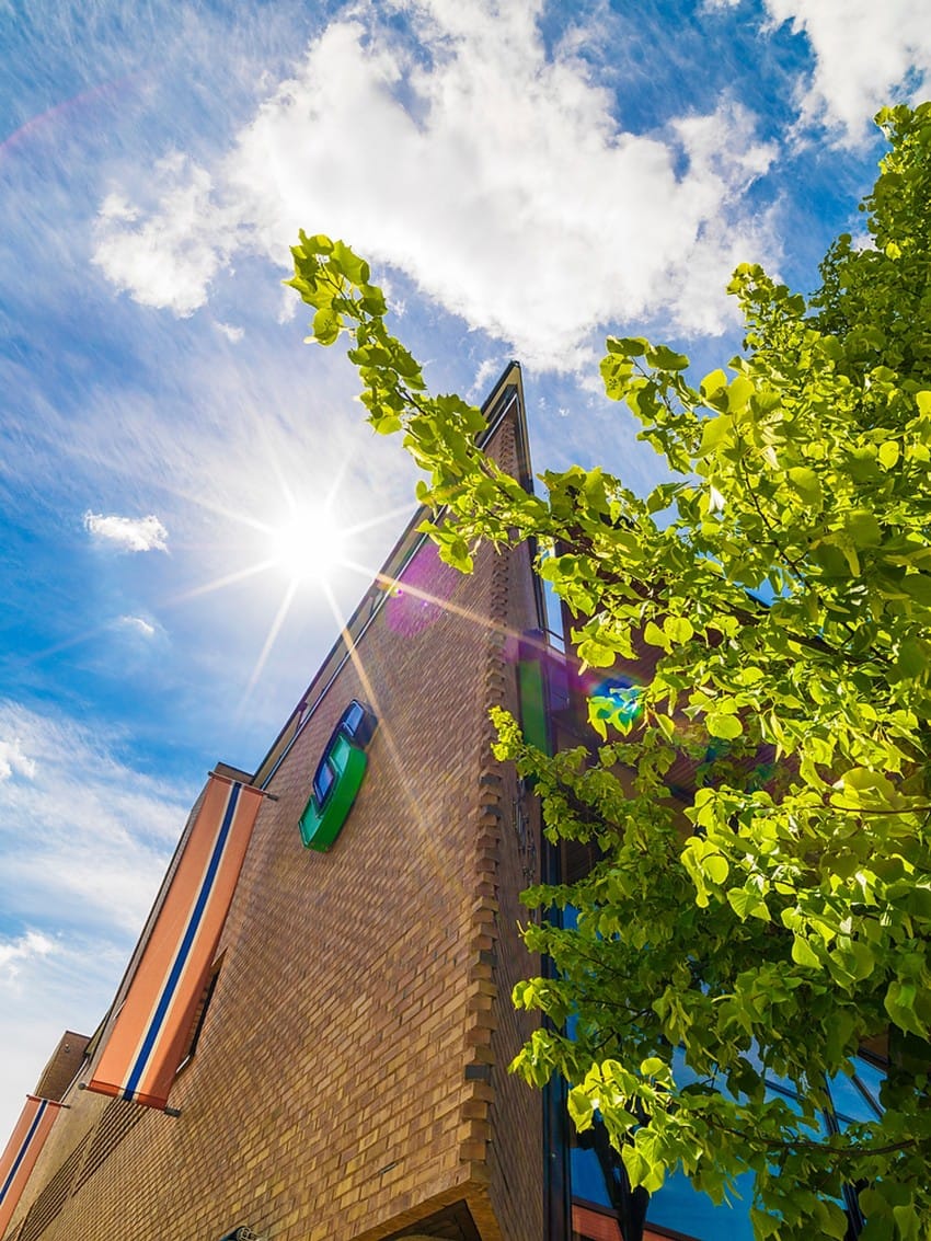 Solen skinner over en murbygning, grønne blader og en klarblå himmel med skyer. Foto: Nathan Lediard, Lediard Foto AS.