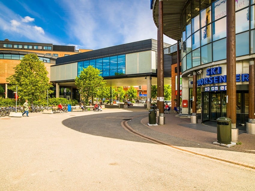Utendørs utsikt over et moderne kjøpesenter med en glassgangvei, trær og sykler parkert i nærheten på en solrik dag. Foto: Nathan Lediard, Lediard Foto AS.