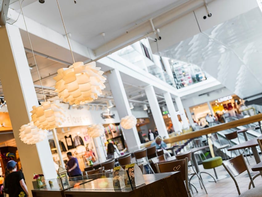 Moderne kjøpesenterinteriør med hvite pendellys, en sittende person og butikker i bakgrunnen. Foto: Nathan Lediard, Lediard Foto AS.