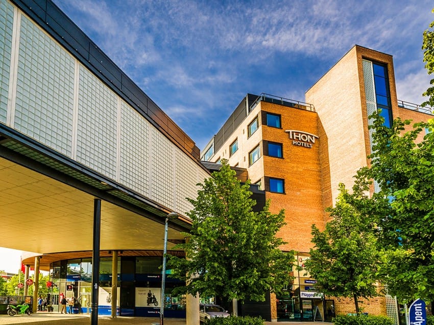 Mursteinsbygning med "Thon Hotel"-skilt, trær i forgrunnen og klarblå himmel over. Foto: Nathan Lediard, Lediard Foto AS.