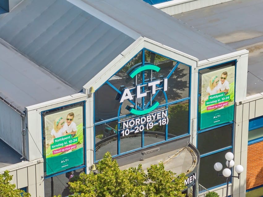 Kjøpesenter med "ALTI NORDBYEN" skilt; to digitale skjermer viser annonser; trær i forgrunnen. Foto: Nathan Lediard, Lediard Foto AS.