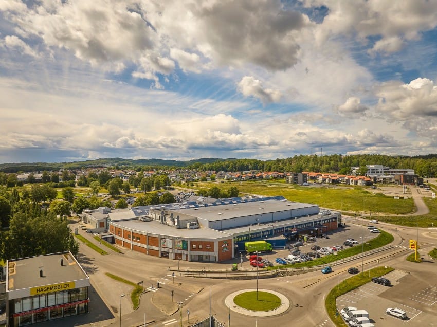 Luftfoto av et forstads kjøpesenter med en parkeringsplass og omkringliggende grøntområder under en delvis overskyet himmel. Foto: Nathan Lediard, Lediard Foto AS.