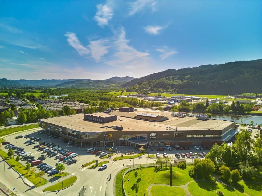 Luftfoto av et stort kjøpesenter med parkeringsplass, omgitt av grønne åser og en elv under en klarblå himmel. Foto: Nathan Lediard, Lediard Foto AS.
