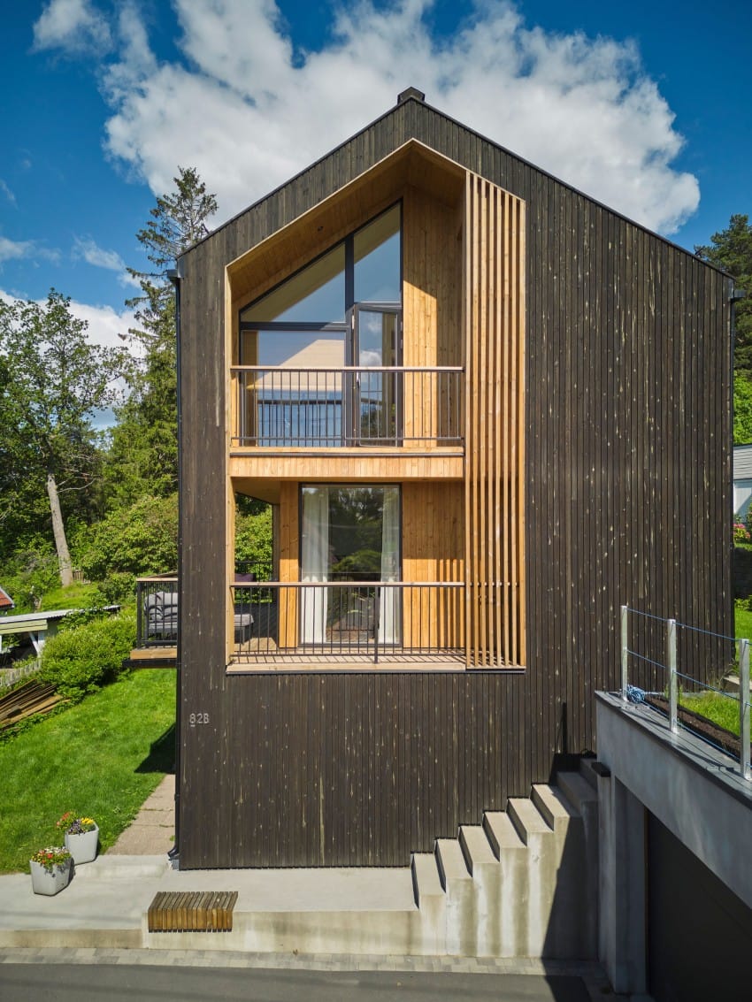 Moderne trehus med store vinduer, skråtak og balkong, omgitt av grønne trær og gress. Foto: Nathan Lediard, Lediard Foto AS.