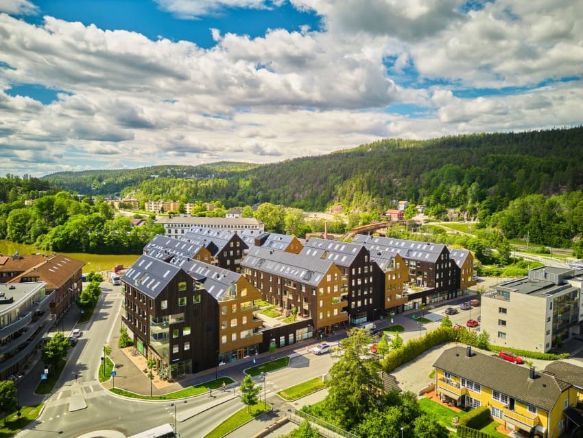 Luftfoto av moderne trebygninger med solcellepaneler omgitt av grøntområder under en delvis overskyet himmel. Foto: Nathan Lediard, Lediard Foto AS.