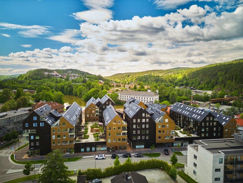 Luftfoto av et moderne boligområde med trehusbebyggelse, omgitt av frodige grøntområder og åser under en delvis overskyet himmel. Foto: Nathan Lediard, Lediard Foto AS.