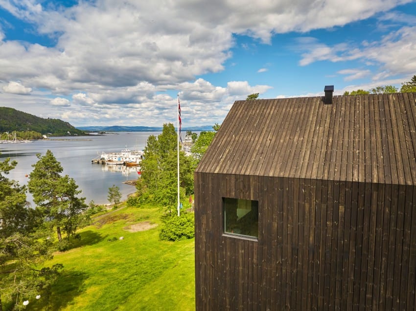 Moderne trehytte med utsikt over en naturskjønn innsjø med båter, trær og en overskyet himmel i bakgrunnen. Foto: Nathan Lediard, Lediard Foto AS.