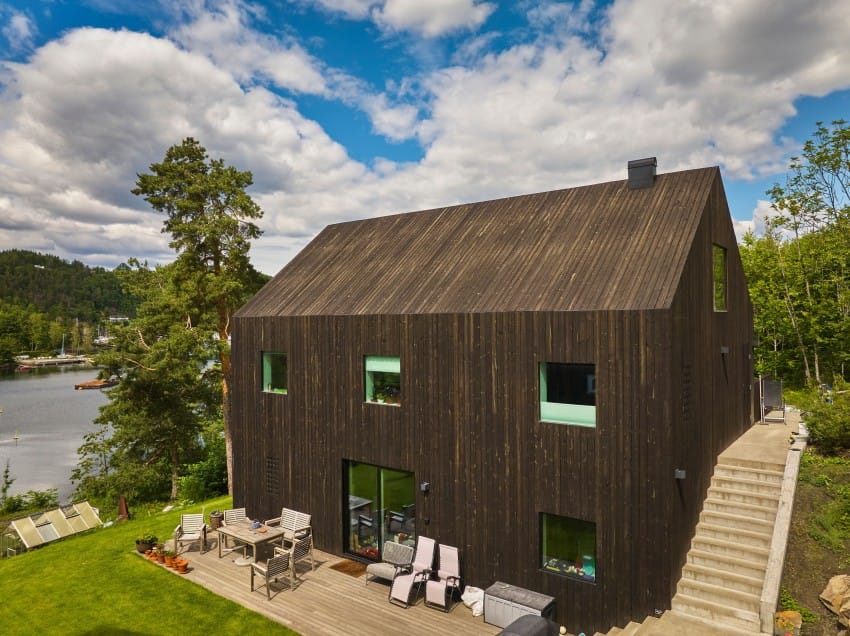 Moderne svart trehus med store vinduer, terrassestoler og omgitt av grøntområder nær en innsjø. Foto: Nathan Lediard, Lediard Foto AS.