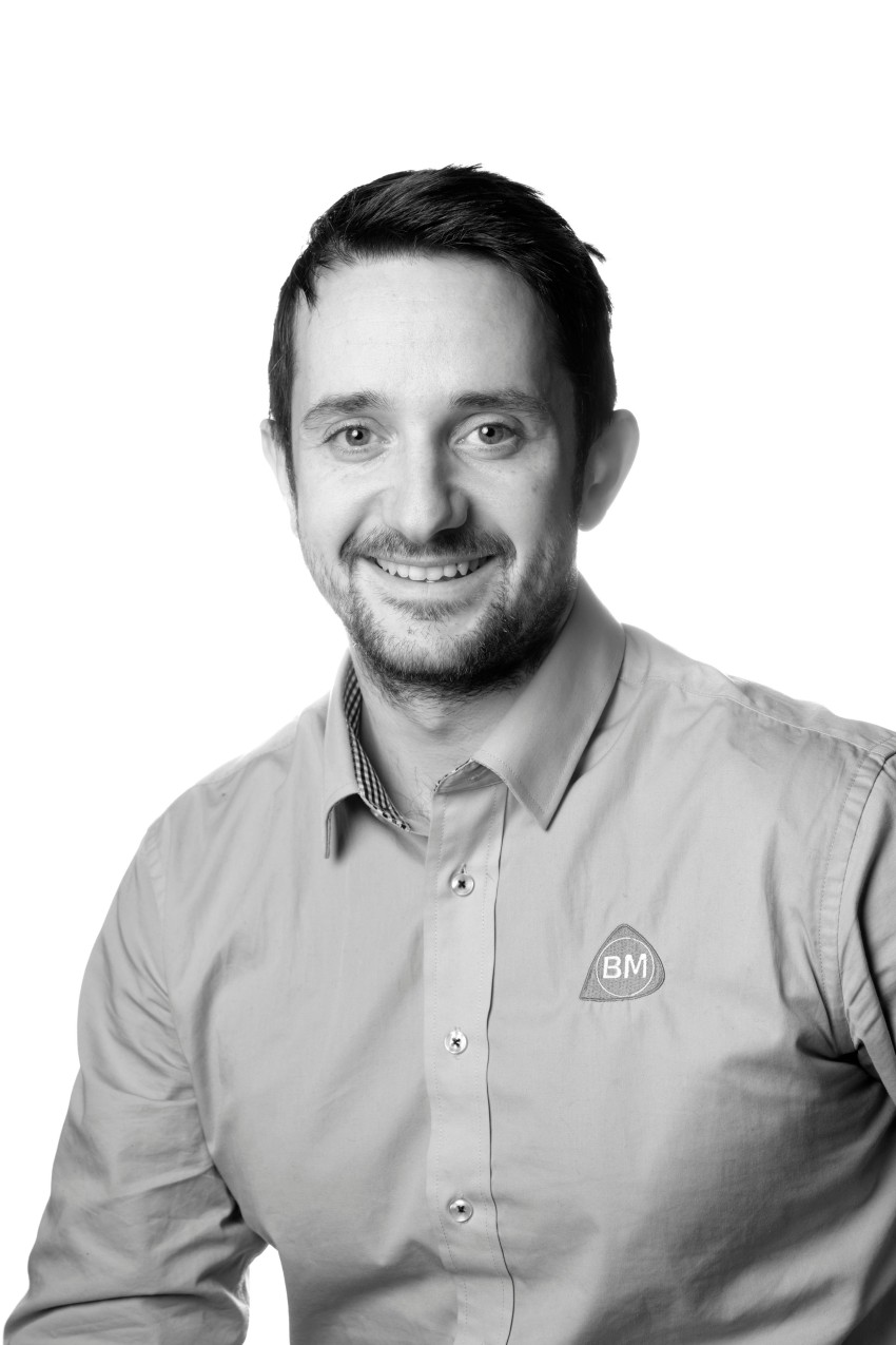 Smilende mann iført en skjorte med en "BM"-logo på hvit bakgrunn. Foto: Nathan Lediard, Lediard Foto AS.