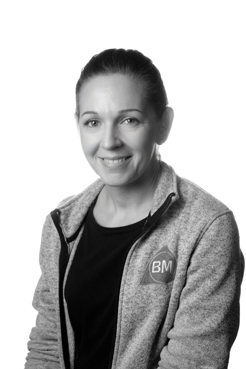 Smilende kvinne i en grå jakke med en "BM"-logo på, poserer mot en hvit bakgrunn. Foto: Nathan Lediard, Lediard Foto AS.