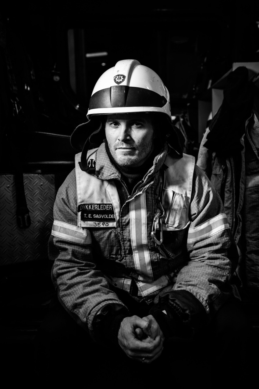 Svart-hvitt-bilde av en brannmann i uniform, som sitter med hendene sammen og ser direkte på kameraet. Foto: Nathan Lediard, Lediard Foto AS.