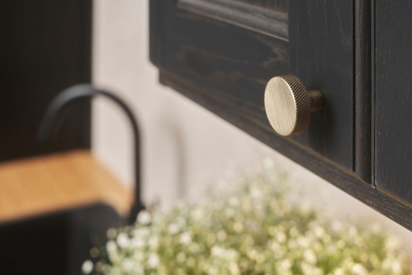 Nærbilde av et mørkt skap med en teksturert gullknott, uskarp kjøkkenvask og blomster i bakgrunnen. Foto: Nathan Lediard, Lediard Foto AS.