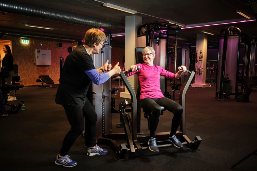 En eldre kvinne trener på en maskin mens en trener gir tommel opp i et treningsstudio. Foto: Nathan Lediard, Lediard Foto AS.
