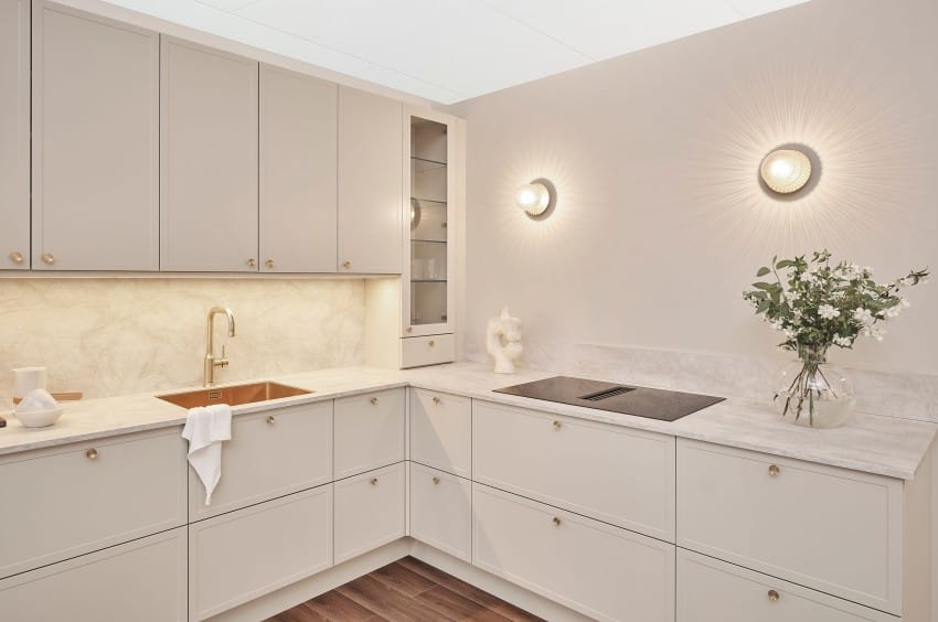 Moderne kjøkken med kremskap, gullarmaturer, vegglamper, induksjonstopp og en vase med blomster på benken. Foto: Nathan Lediard, Lediard Foto AS.