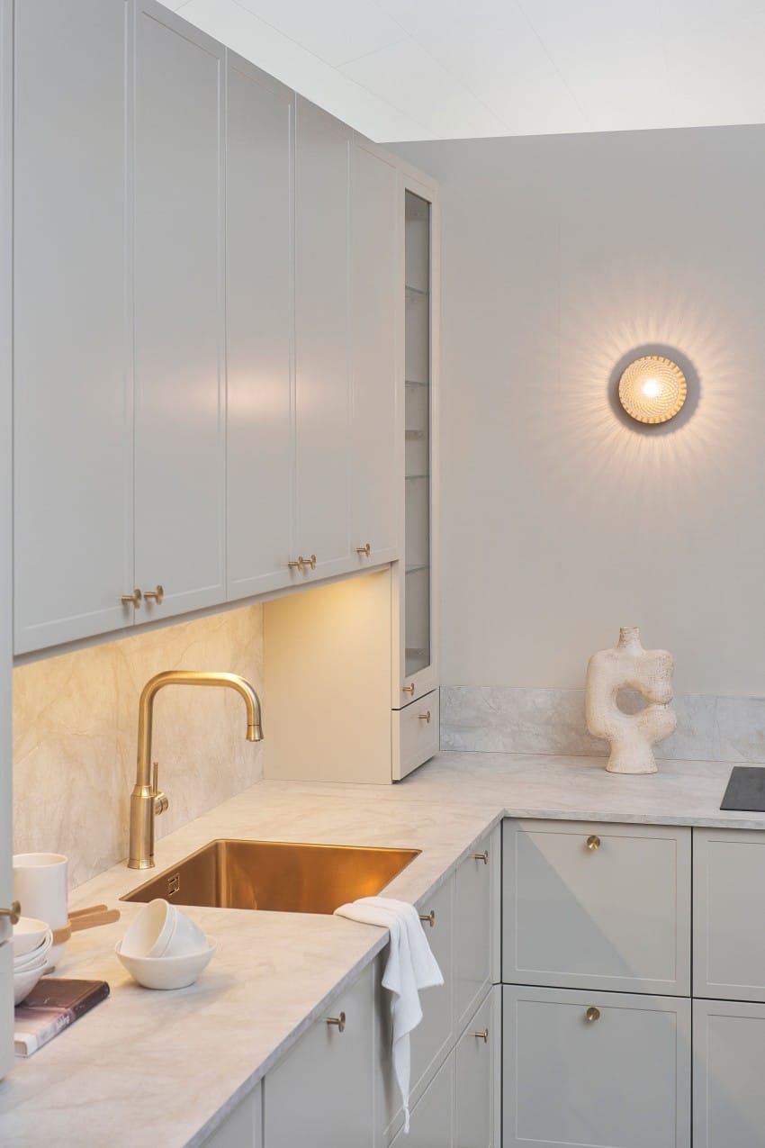 Moderne kjøkken med lysegrå skap, benkeplater i marmor, gullkran, veggskulptur og sirkulær vegglampe. Foto: Nathan Lediard, Lediard Foto AS.