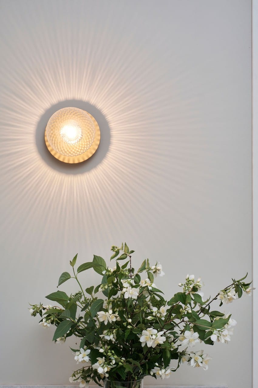 Rund vegglampe som kaster lysmønstre over hvite blomster og grønne blader mot en blek vegg. Foto: Nathan Lediard, Lediard Foto AS.
