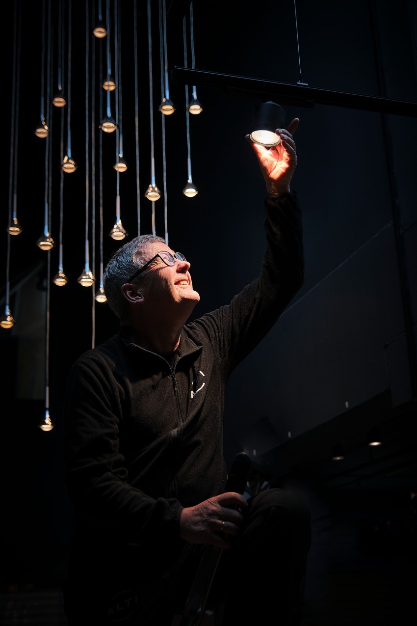 Person som justerer et hengende lys i et svakt opplyst rom, omgitt av flere små lys. Foto: Nathan Lediard, Lediard Foto AS.