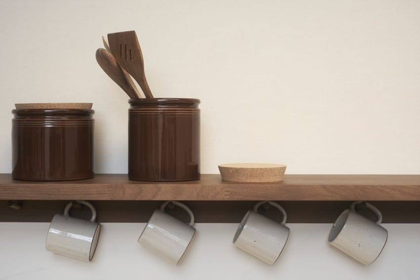 Trehylle med to brune krukker, kokekar, et bord og fire hengende hvite krus mot en kremvegg. Foto: Nathan Lediard, Lediard Foto AS.