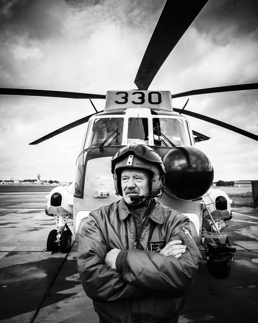Pilot i hjelm og jakke står selvsikkert foran et helikopter på asfalten, nummerert 330, i gråtoner. Foto: Nathan Lediard, Lediard Foto AS.