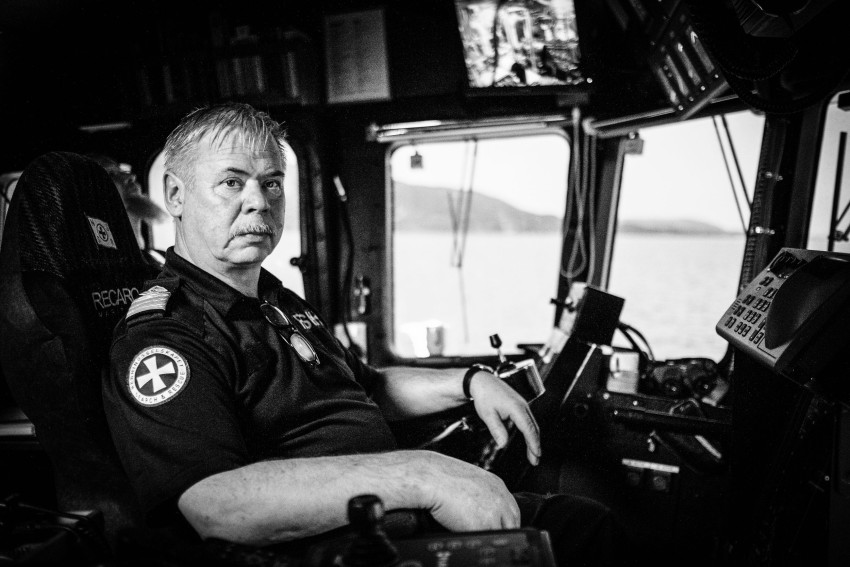 Svart-hvitt bilde av en kaptein som sitter i et skips kontrollrom og ser intenst mot kameraet. Foto: Nathan Lediard, Lediard Foto AS.