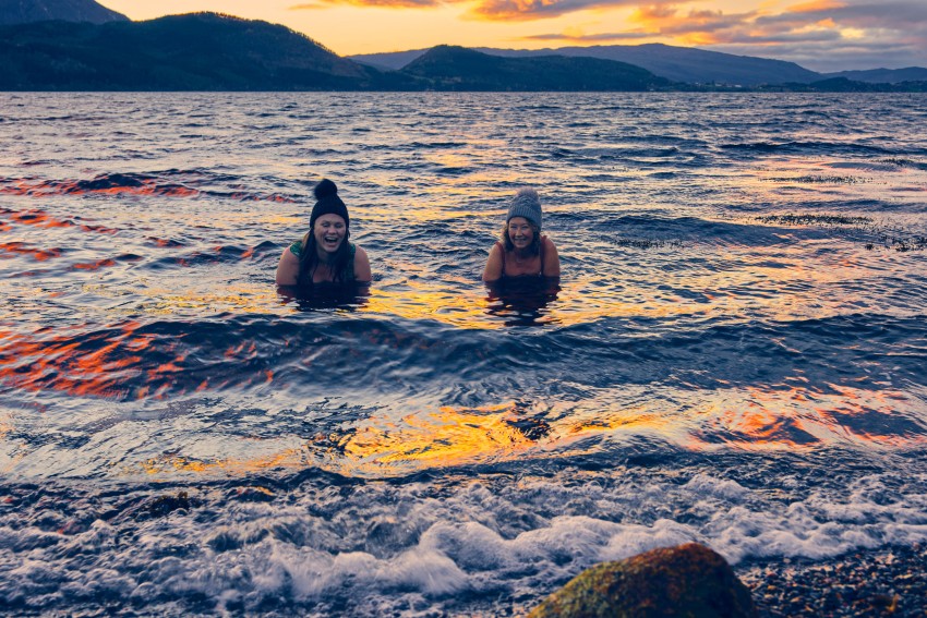 To personer svømmer i en innsjø ved solnedgang, iført luer, med fjell i bakgrunnen. Foto: Nathan Lediard, Lediard Foto AS.