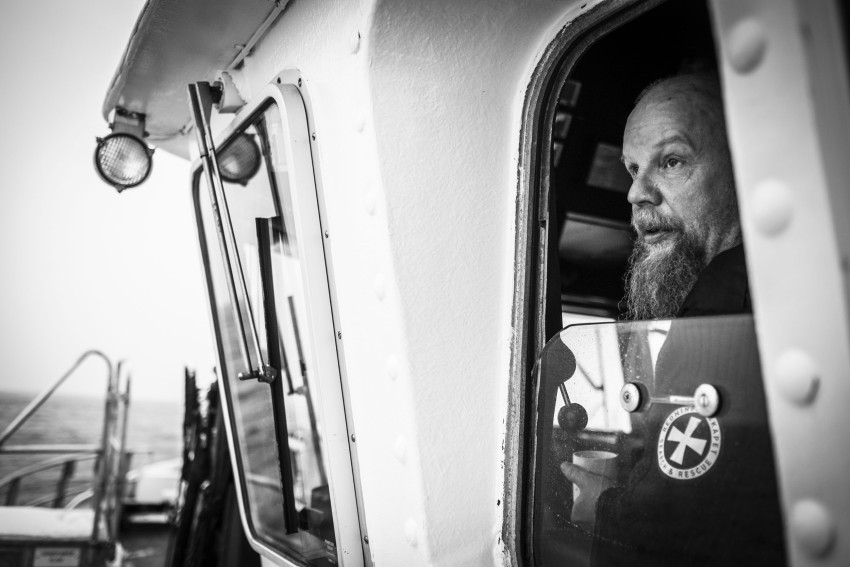 Skjeggete mann med joystick som fører en båt, sett gjennom et vindu, i et svart-hvitt bilde. Foto: Nathan Lediard, Lediard Foto AS.