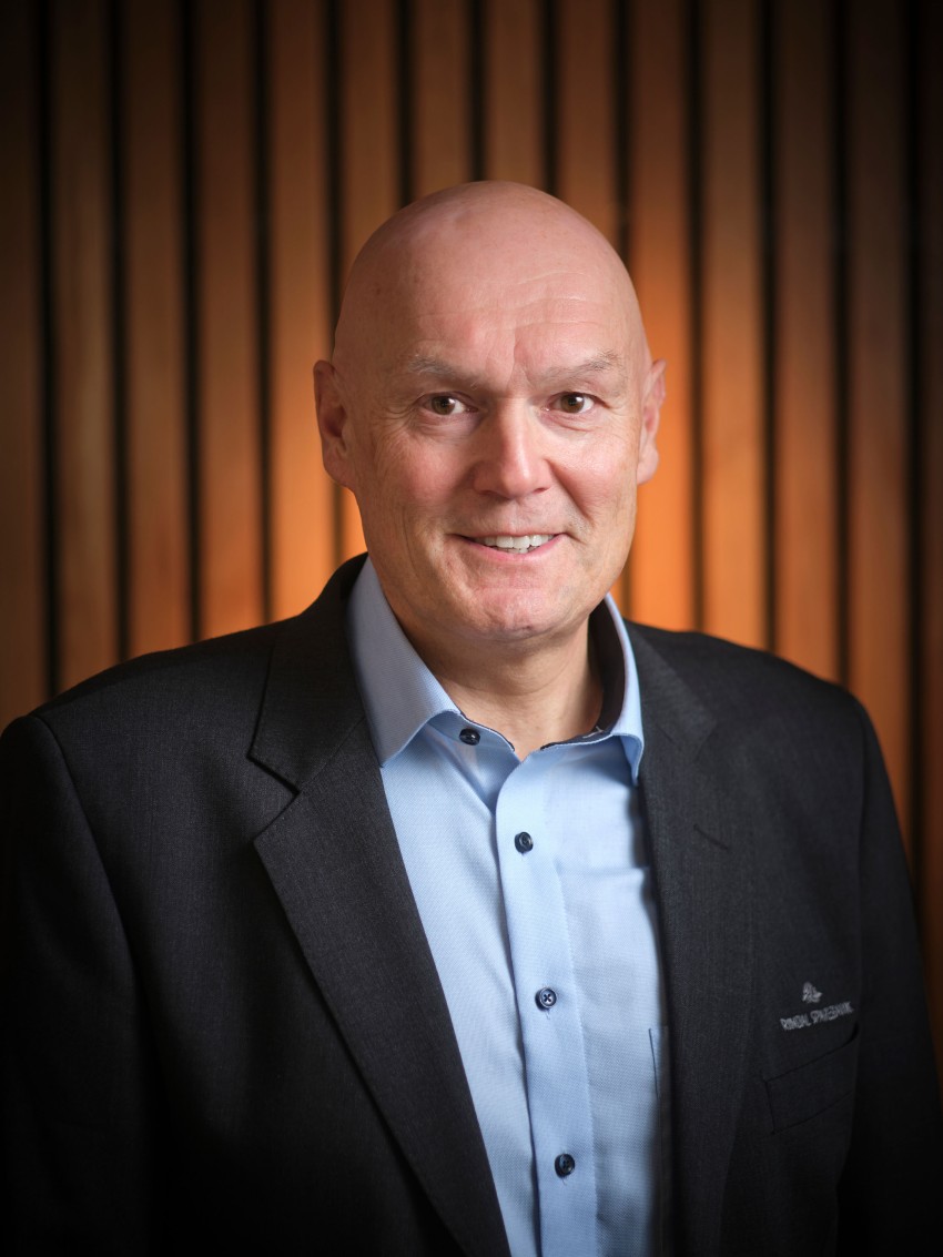 En smilende mann i dress med skallet hode står mot en trelistbakgrunn. Foto: Nathan Lediard, Lediard Foto AS.