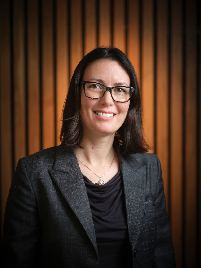 Smilende person med briller i en grå dress står mot en bakgrunn av trepanel. Foto: Nathan Lediard, Lediard Foto AS.