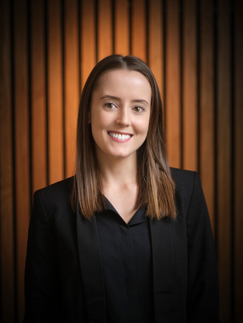 Smilende kvinne med rett, brunt hår iført en svart blazer, stående mot en trelistbakgrunn. Foto: Nathan Lediard, Lediard Foto AS.