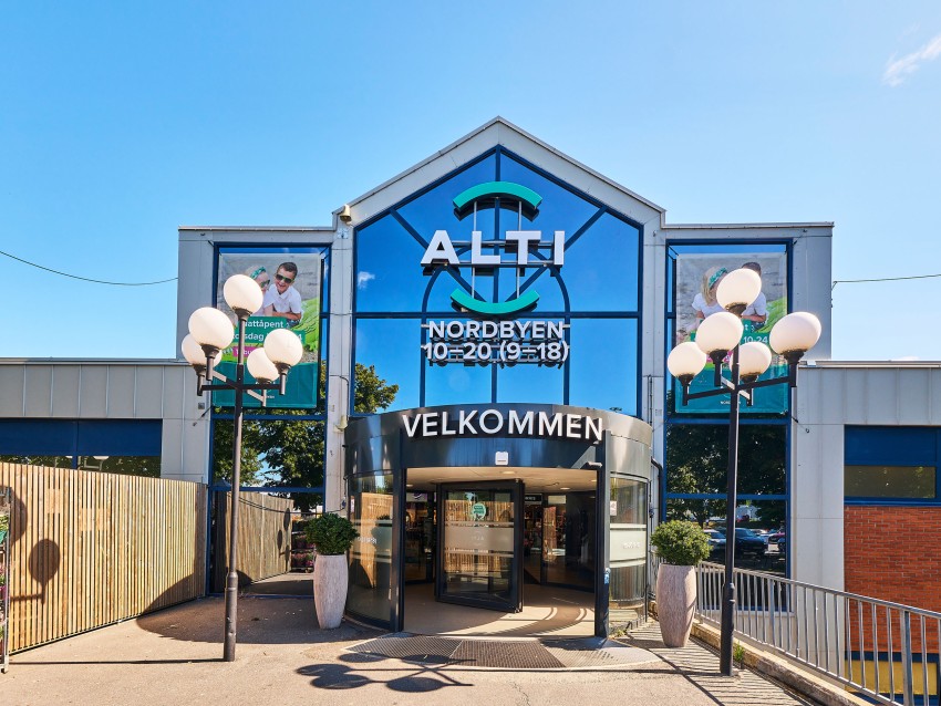 Kjøpesenterinngang med glassfasade, skilt "Alti Nordbyen", og svingdører. Klar blå himmel. Foto: Nathan Lediard, Lediard Foto AS.