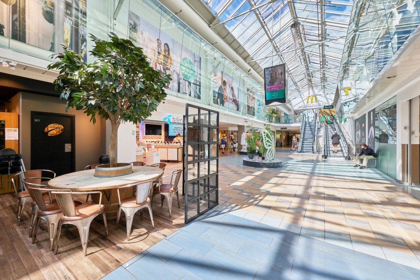 Romslig kjøpesenter med takvinduer, sittegruppe, rulletrapper og butikker i sterkt naturlig lys. Foto: Nathan Lediard, Lediard Foto AS.