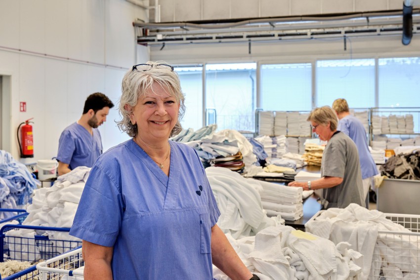 Eldre kvinne smilende i et vaskerom iført blå uniform, mens andre sorterer klær i bakgrunnen. Foto: Nathan Lediard, Lediard Foto AS.