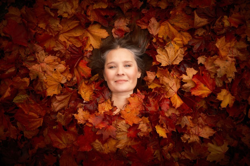 En kvinne smilende, liggende på ryggen midt i en haug med livlige høstløv. Foto: Nathan Lediard, Lediard Foto AS.